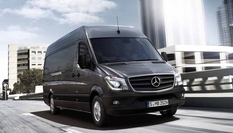 Large, black Mercedes Benz rental van driving through a city, past modern, office buildings. 